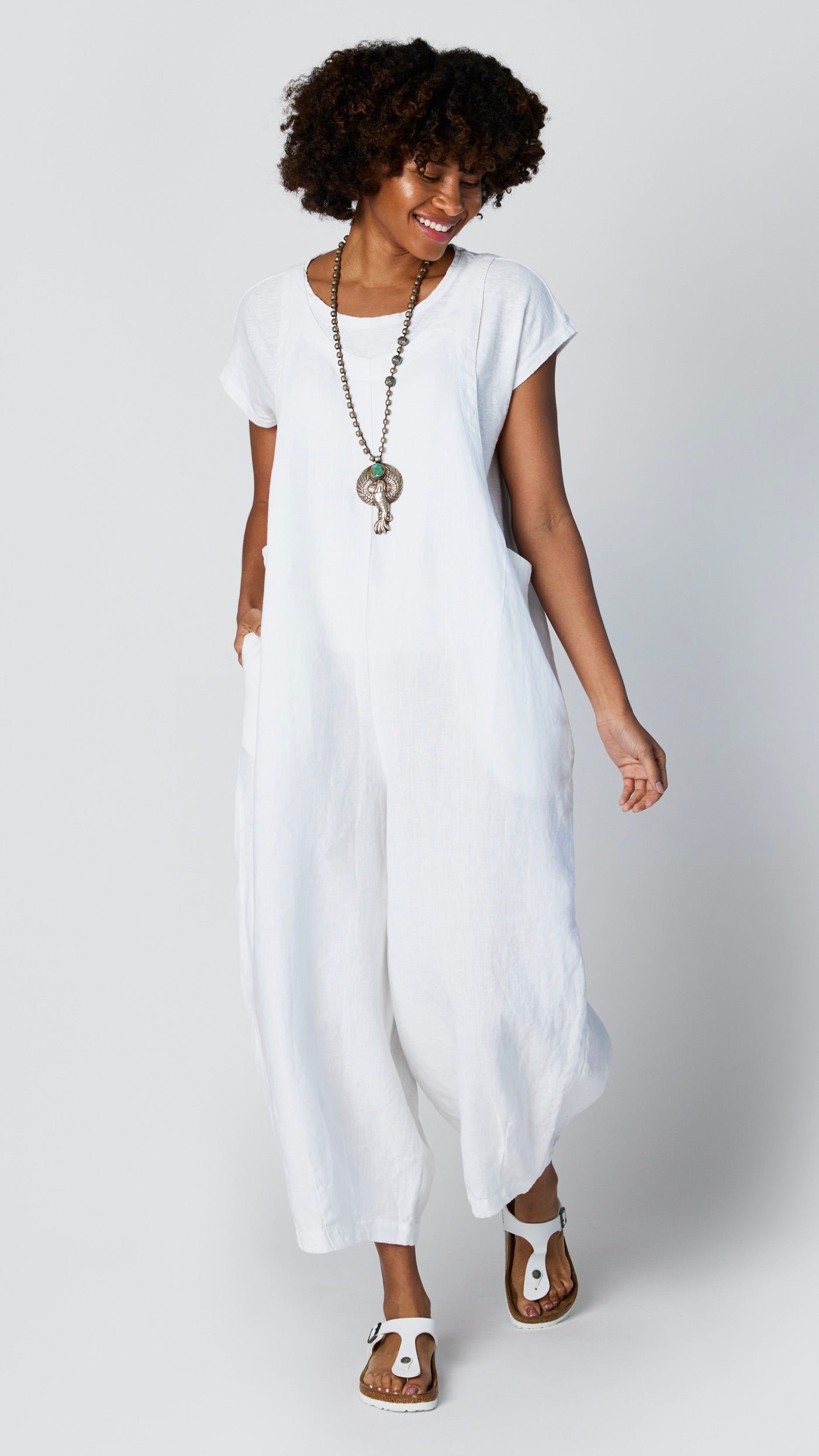Model wearing white linen overall jumpsuit with wide cropped dart leg, white 3/4 sleeve bias cut tee, and white sandals.