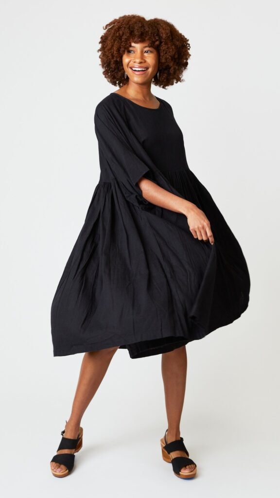 Model wearing oversized black cotton dress with raglan sleeves and waist-gathered skirt, and black leather sandals.