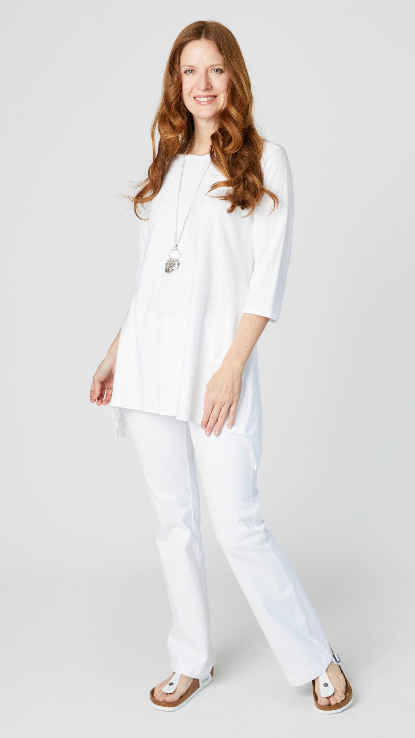 Model wearing white 3/4 sleeve high-low tunic with white bootcut jeans, long silver pendant necklace, and white sandals