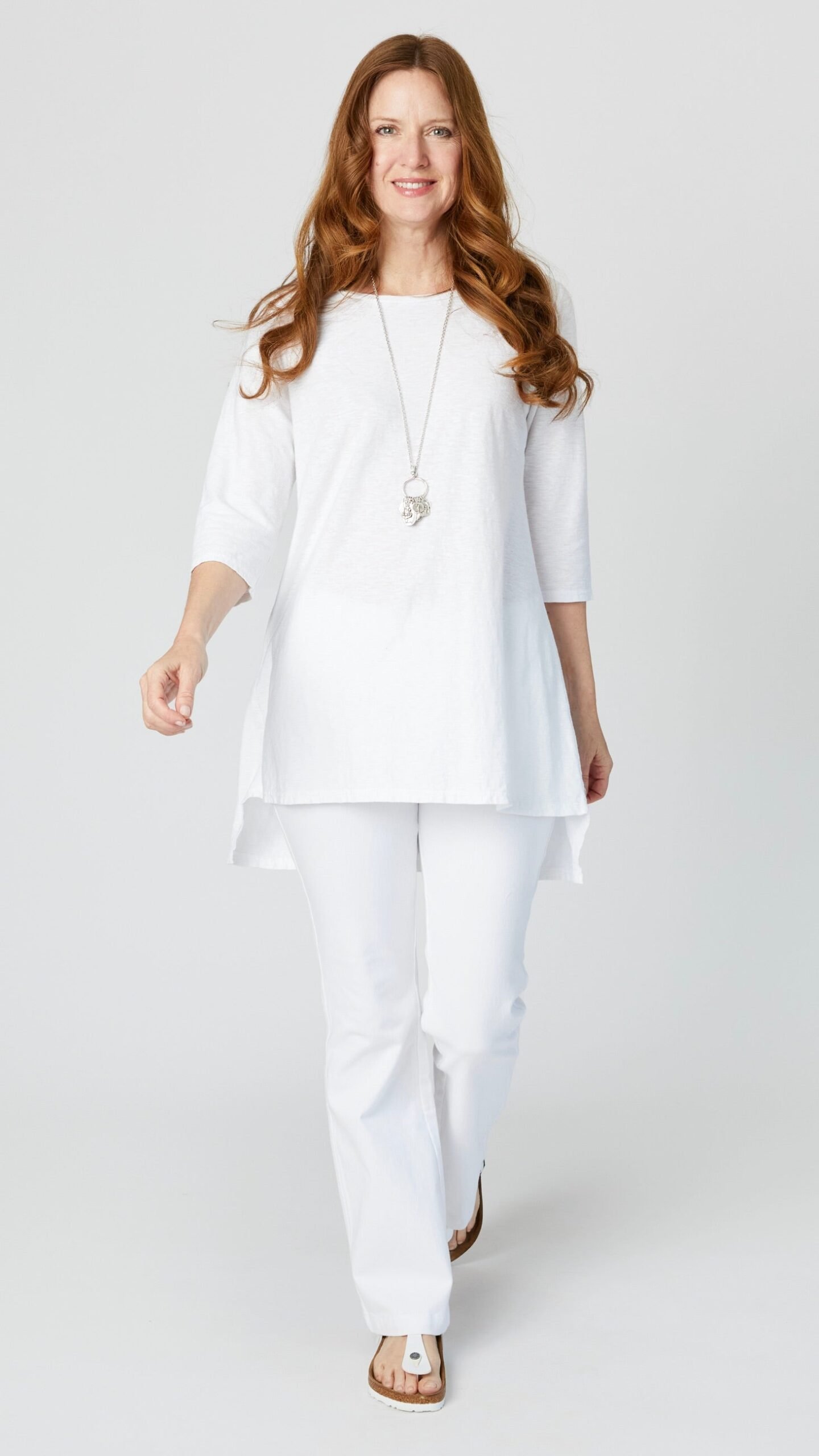 Model wearing white 3/4 sleeve high-low tunic with white bootcut jeans, long silver pendant necklace, and white sandals