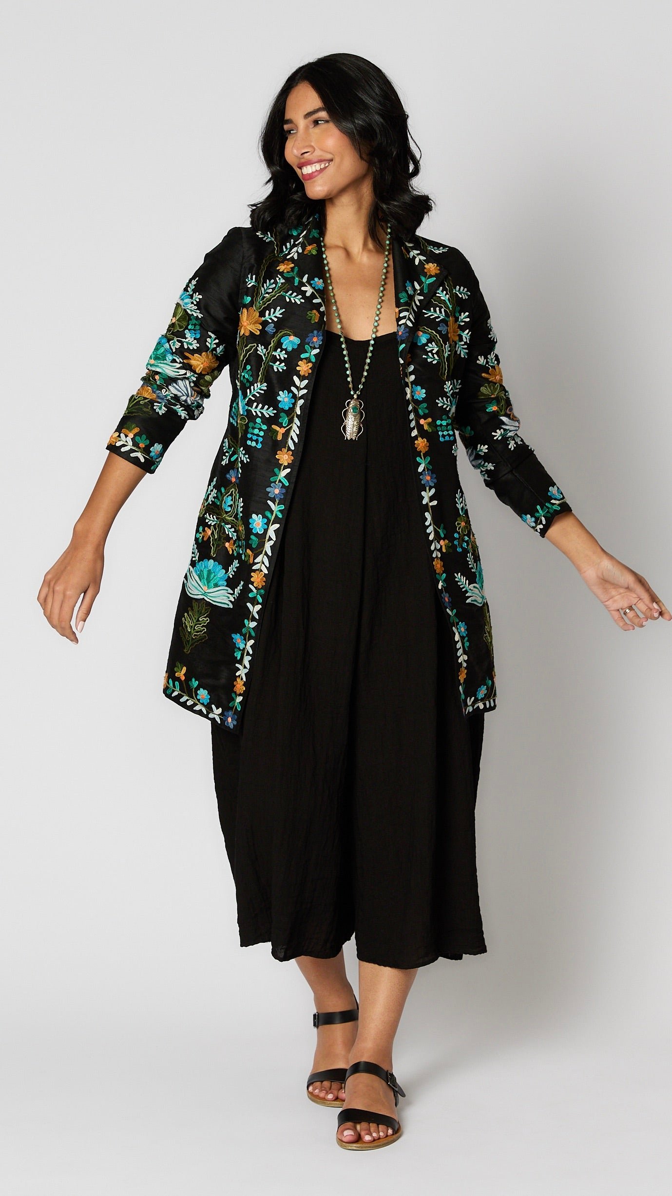 Model wearing black silk duster embroidered with bright blue, yellow and green tones over black rayon romper and black leather sandals.