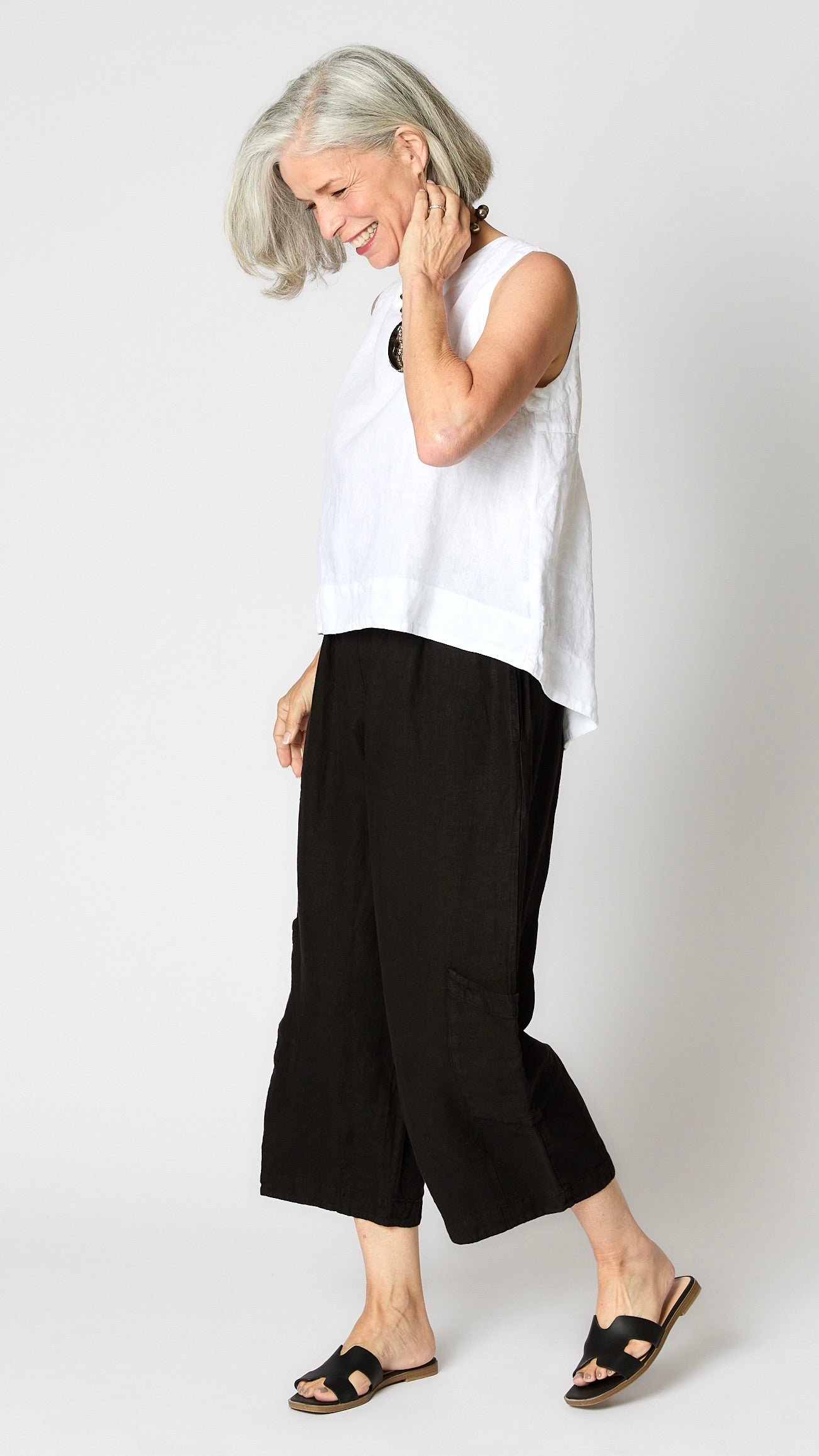 Model wearing white linen hi-lo tank top with dyed bone moon pendant necklace, black cropped linen pants with ankle darts and patch pockets, and black sandals.
