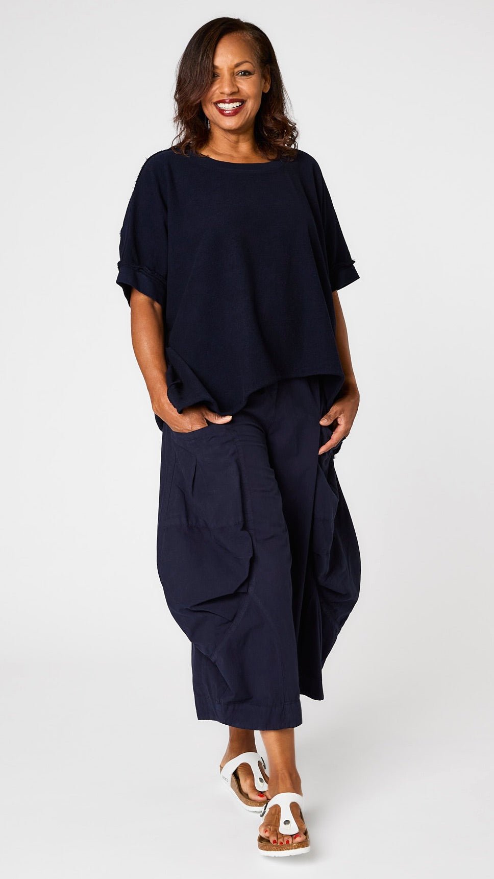 Model wearing indigo short sleeve pullover vest with oversized drop shoulder, crew neck, and curved hi-lo hemline over indigo double-pocket pant, and white sandals.