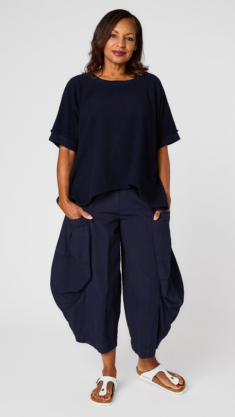 Model wearing indigo short sleeve pullover vest with oversized drop shoulder, crew neck, and curved hi-lo hemline over indigo double-pocket pant, and white sandals.