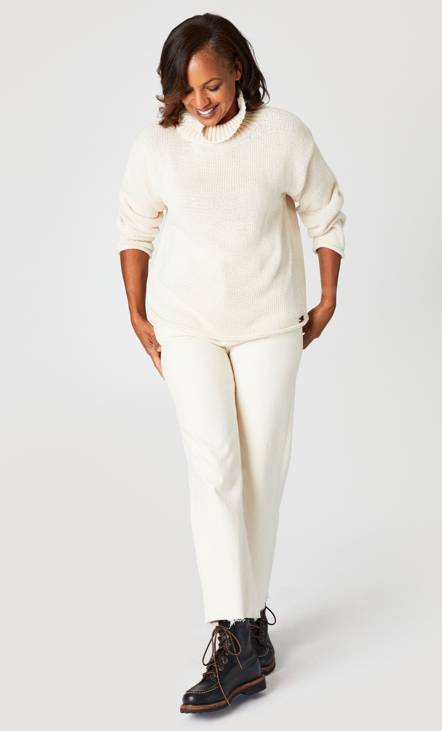 Model wearing hip-length hand knit cotton turtleneck sweater, and wide leg ankle length denim jeans with raw hem in ecru.