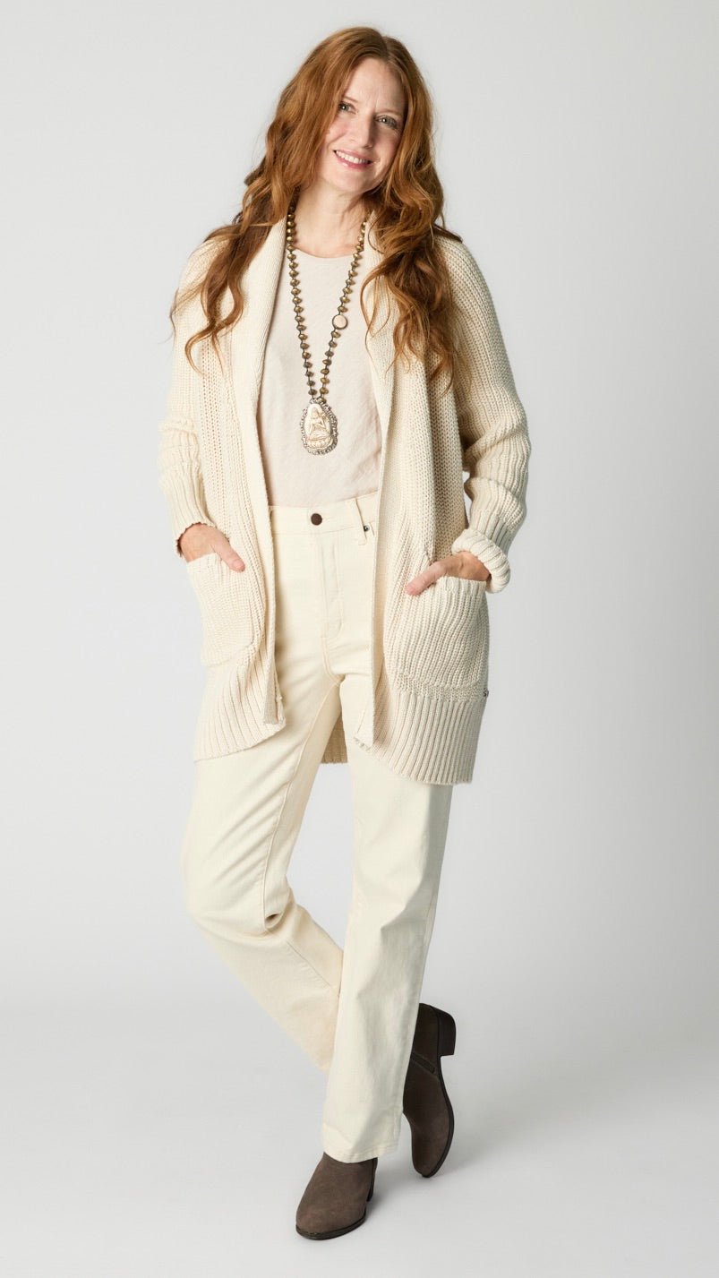 Model wearing cream cotton handknit oversized cardigan (without buttons) over natural bias top, ecru straight leg denim pant, and taupe suede boot.