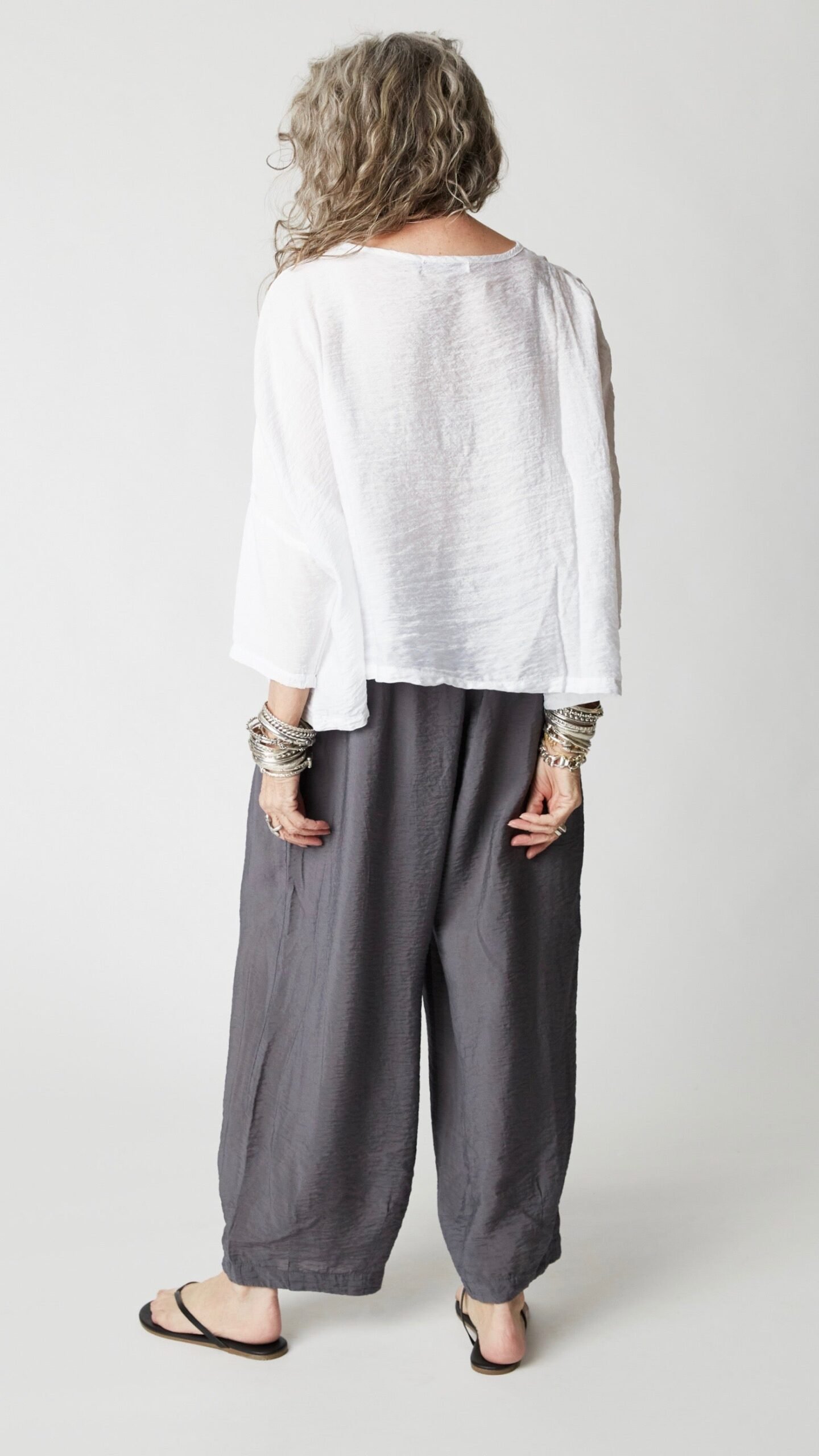 Model wearing cropped white rayon top with high round neckline, dropped shoulders and ¾ sleeves, gray cropped dart pants, silver bangles, and black flip-flops.