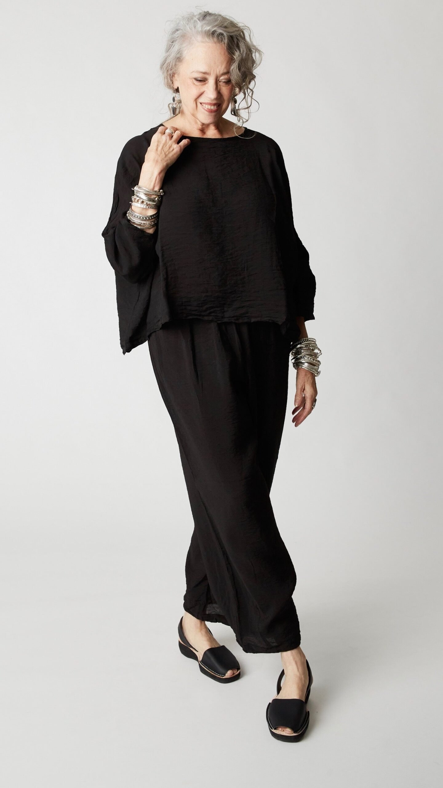 Model wearing cropped black rayon top with high round neckline, dropped shoulders and ¾ sleeves, black cropped dart pants, silver bangles, and black open-toed flats.
